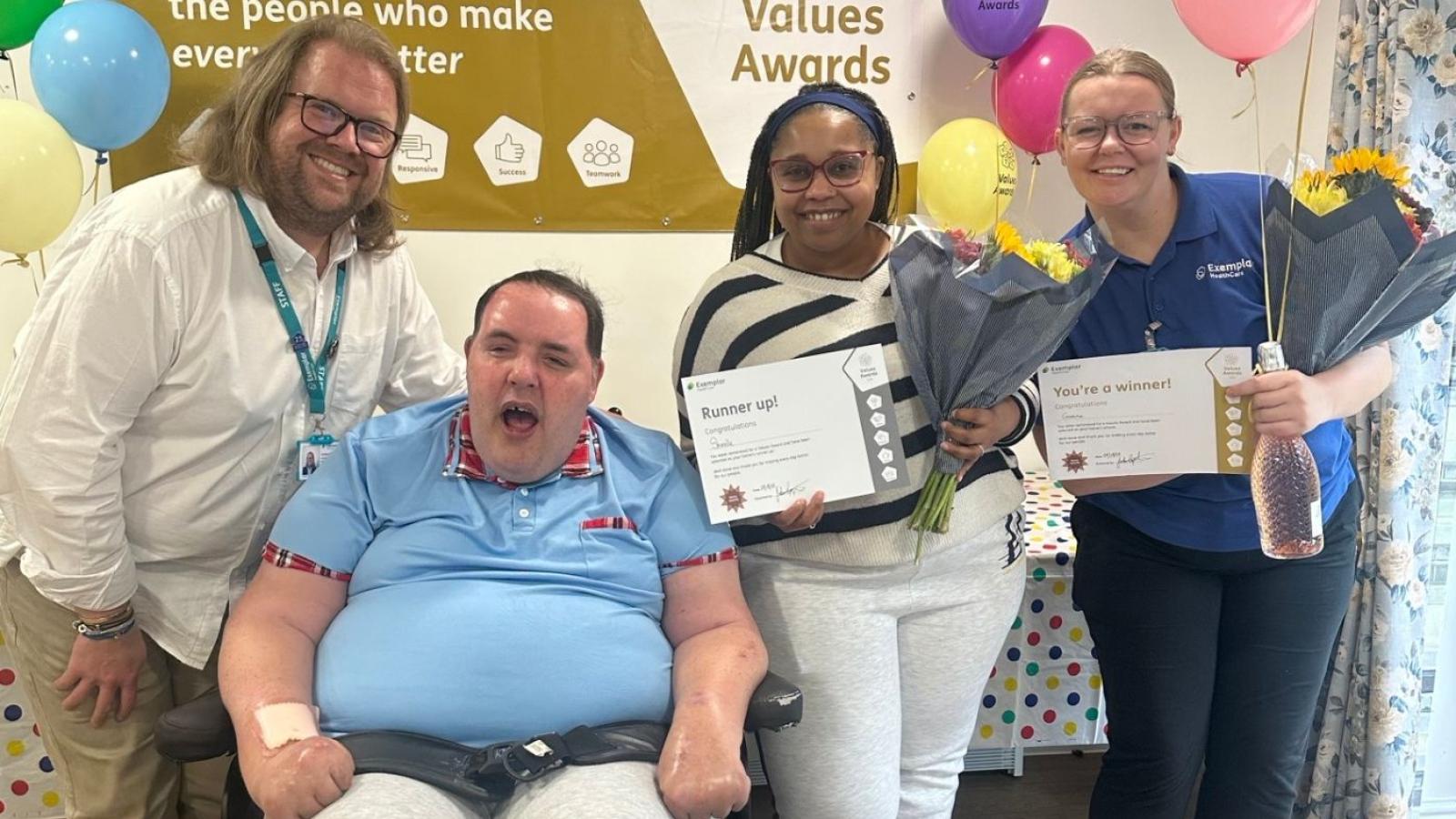 Four people, one in a wheelchair, posing for a photo. Two are holding certificates and flowers