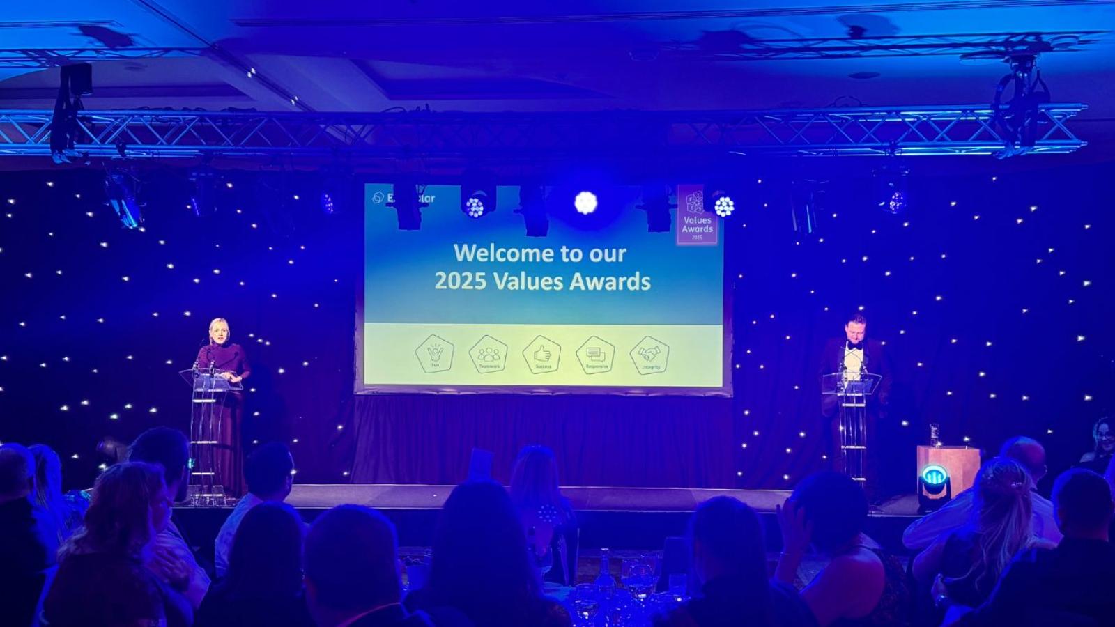 A screen saying 'Values Awards 2025' with two presenters stood either side, in front of a room full of people