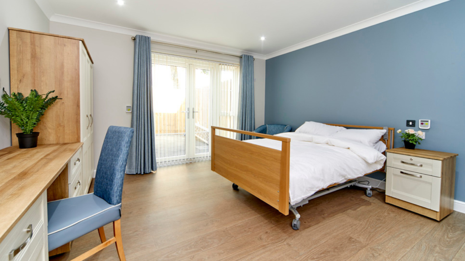 A bedroom with dressing table, chair, wardrobe, double bed, drawers and double patio doors leading to a garden