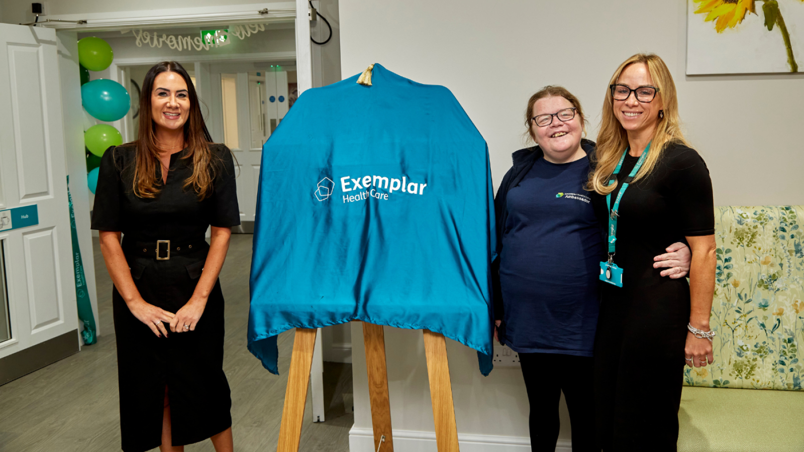 Three people stand beside an easel with a drape over smiling