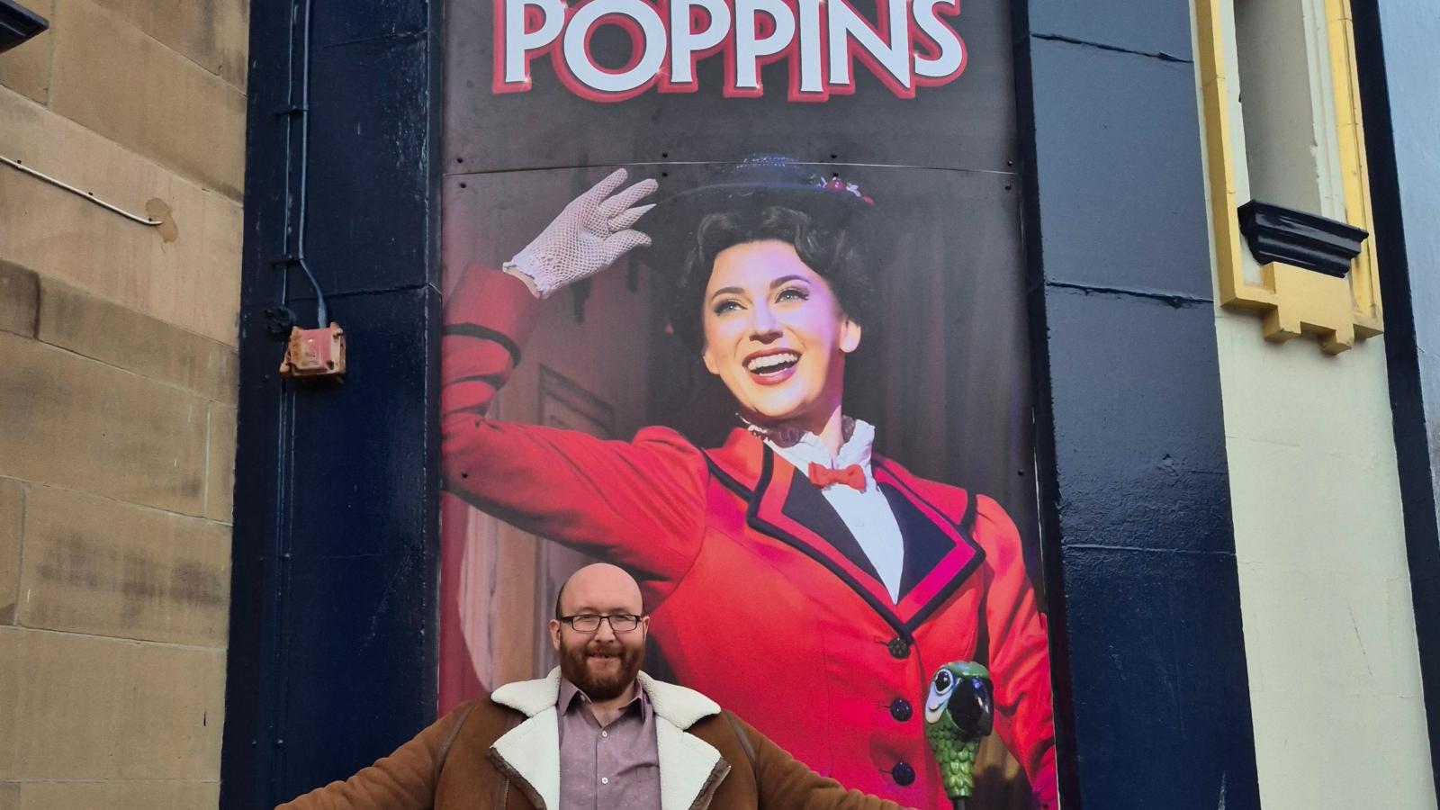 Smiling man stood infront of a promotional poster for Mary Poppins.