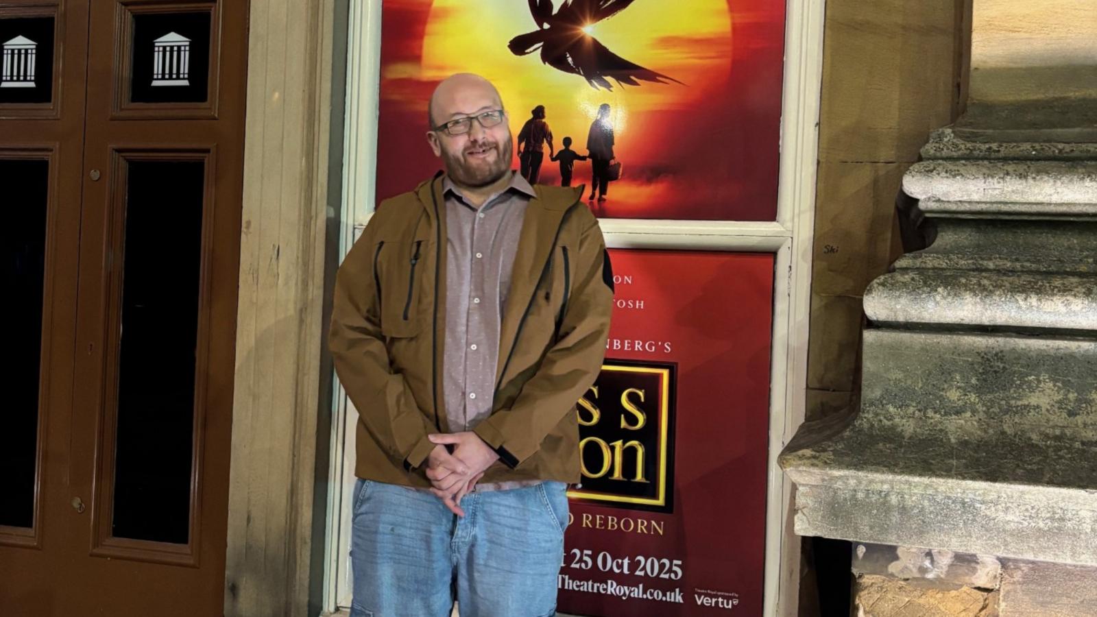 Smiling man stood infront of a promotional poster for Miss Saigon.