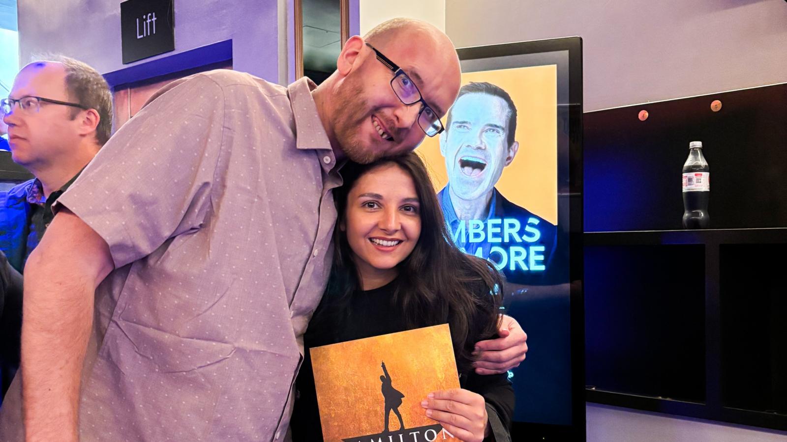 Two people smiling and posing together at a 'Hamilton' musical event, holding a program booklet, with a promotional poster visible in the background.