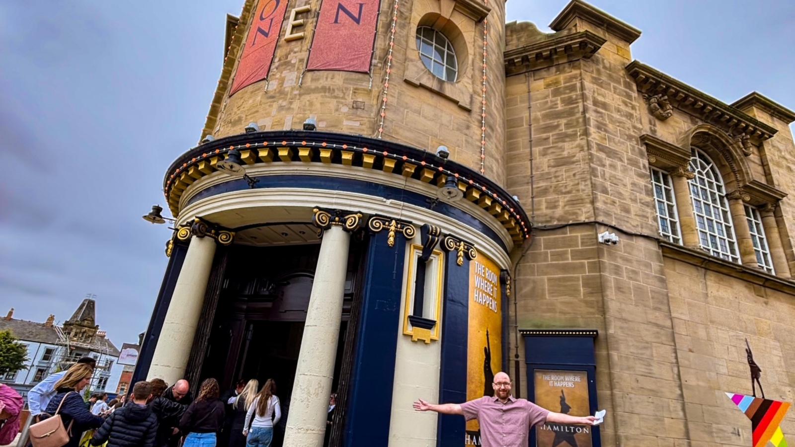 Smiling man stood infront of a promotional poster for Hamilton.