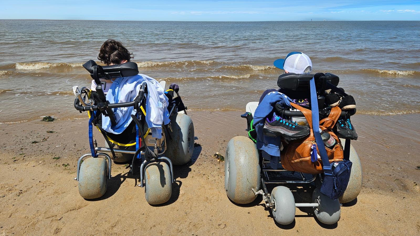 Two individuals using specialized beach wheelchairs are facing the sea on a sandy beach under a clear blue sky.