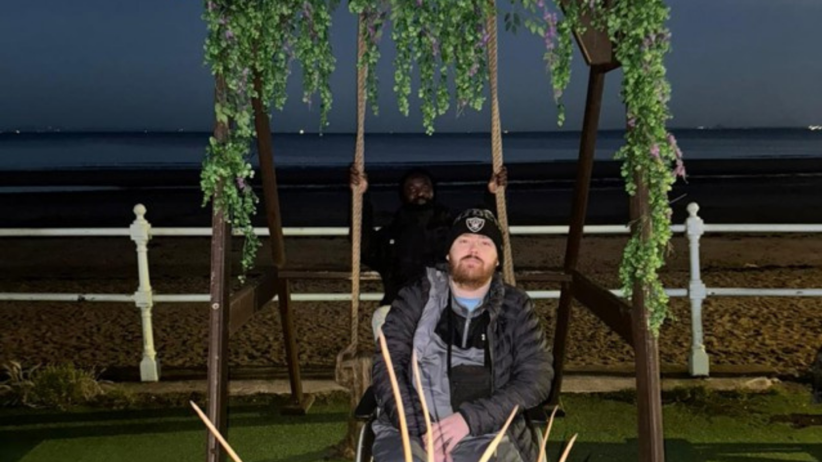 Person in a wheelchair sitting under a rustic wooden sign that reads "BOATHOUSE," adorned with hanging green plants, near a railing overlooking the sea at dusk.