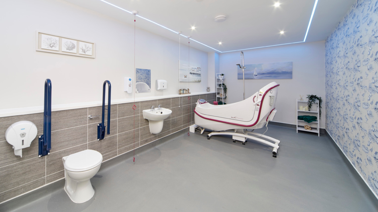 A modern, spacious bathroom with white walls and grey flooring. Features a toilet with blue grab bars, a mounted sink, and a high-tech bathtub. The ambiance is clean and calming.
