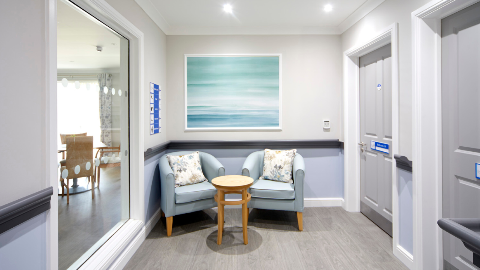 Cozy corner with two light blue armchairs, a small table, and a large ocean-themed painting. Soft, calming tone with grey doors and natural light.