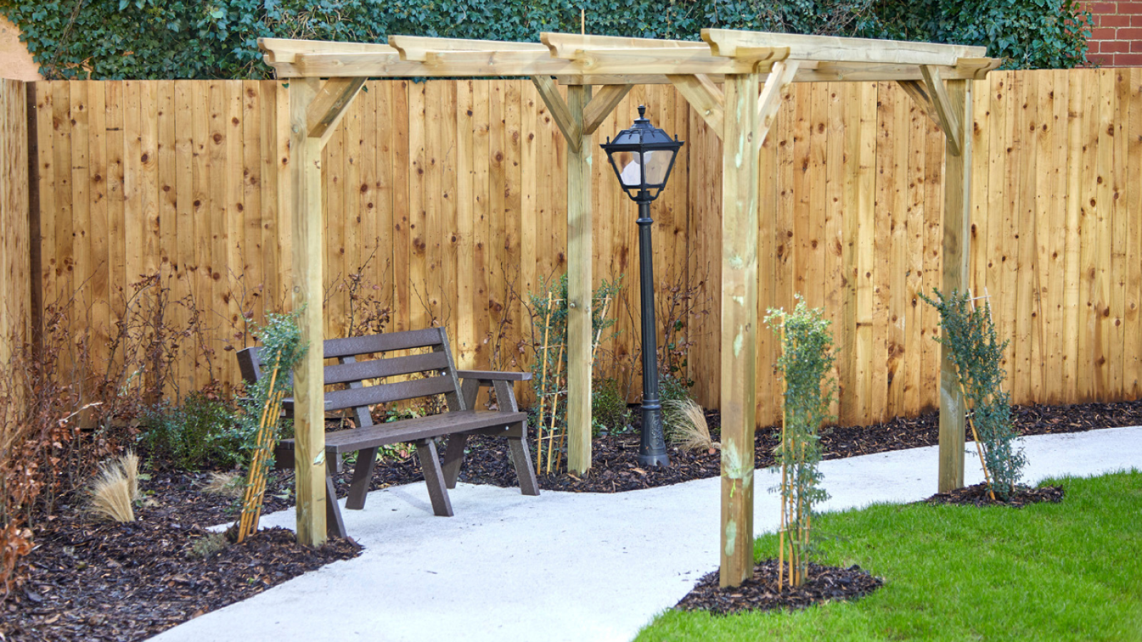 A wooden pergola stands over a curved white pathway next to a bench and vintage lamp post, surrounded by green grass and bordered by a wooden fence.