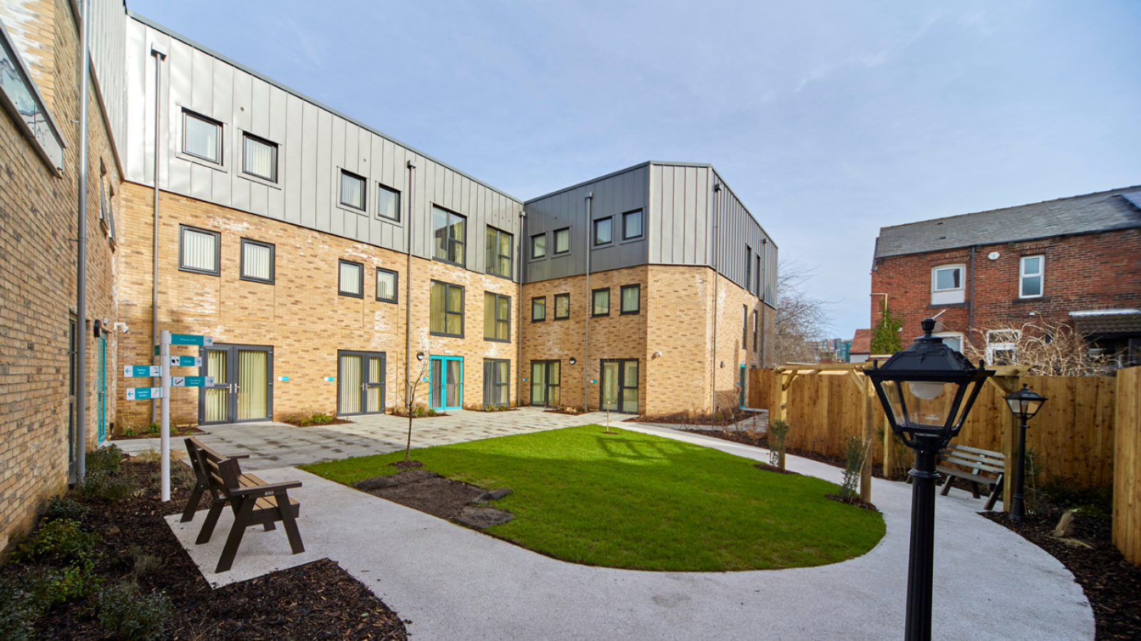 Modern brick building with metal roof surrounding a small courtyard. The courtyard features a neatly paved path, green lawn, benches, and a vintage-style lamp post under a clear blue sky.