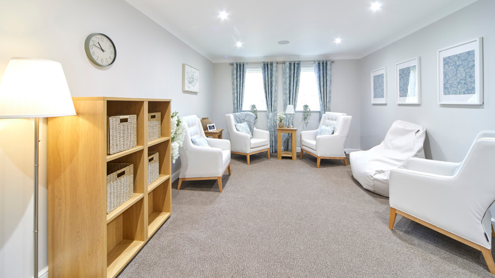 A cozy living room features white armchairs, a white beanbag, and wooden accents. Soft blue curtains and framed art add calm. A lamp and shelf with baskets complete the serene vibe.