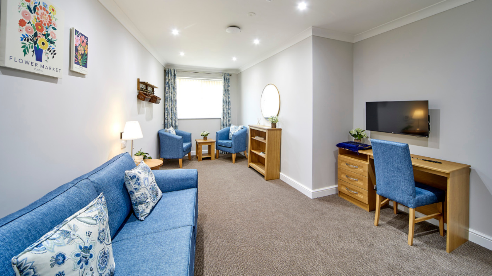 Cozy living space with blue sofas, floral cushions, and a wall-mounted TV. A wooden desk and armchairs complement the neutral tones and bright ambiance.