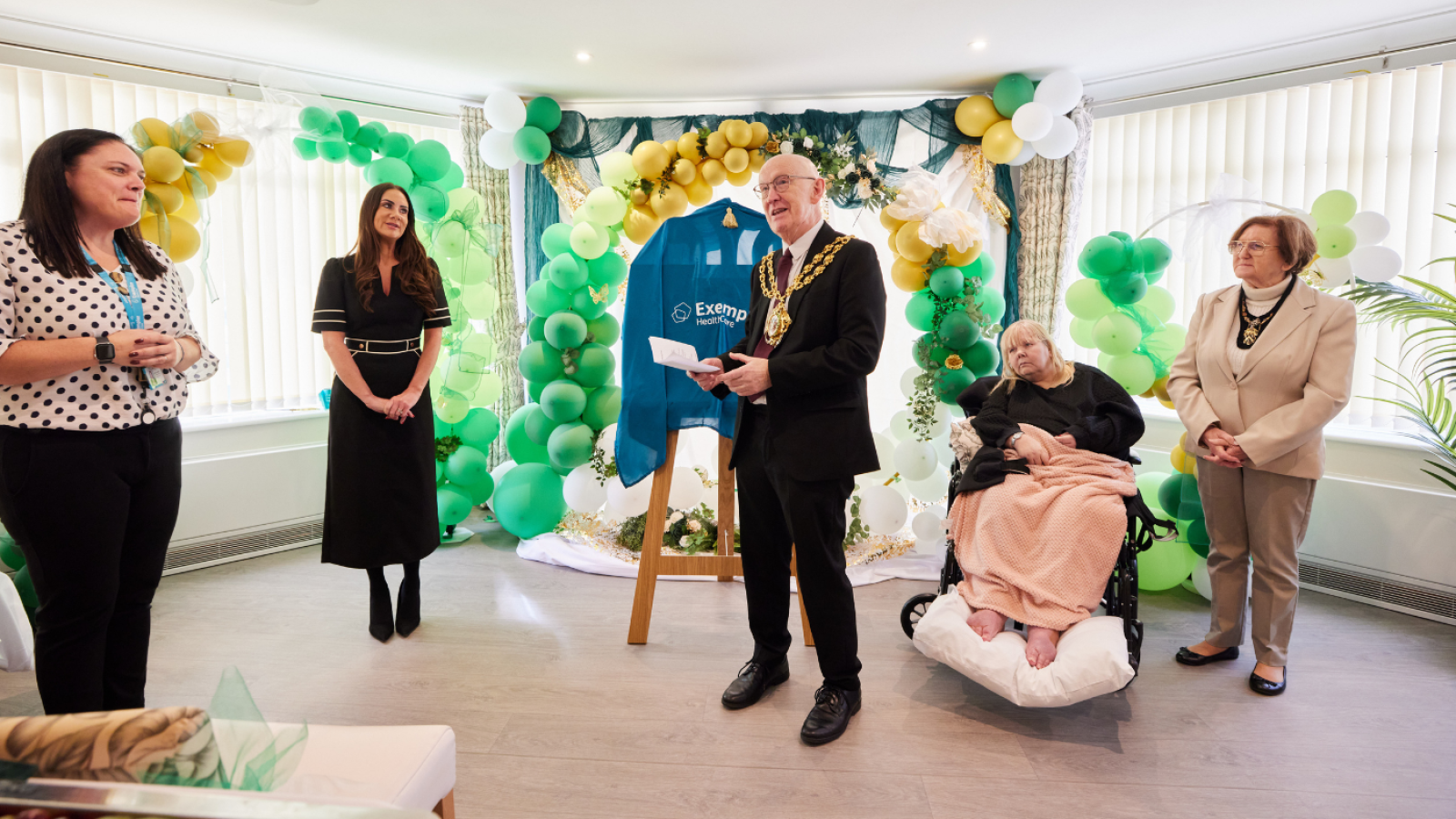A group of five people stand in a bright room decorated with green, gold, and white balloons. A man wearing a chain of office speaks by a display. A woman in a wheelchair is nearby with a pink blanket, alongside two women standing. The mood is festive and formal.