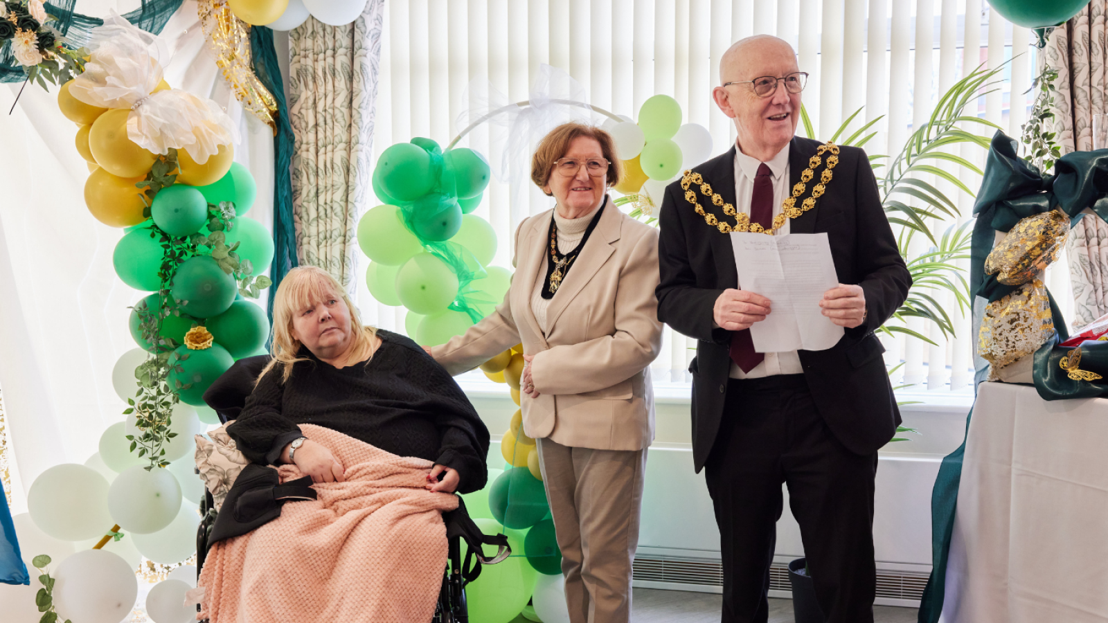A woman in a wheelchair with a pink blanket is beside two smiling older adults in formal attire. They stand near green and yellow balloon decorations.