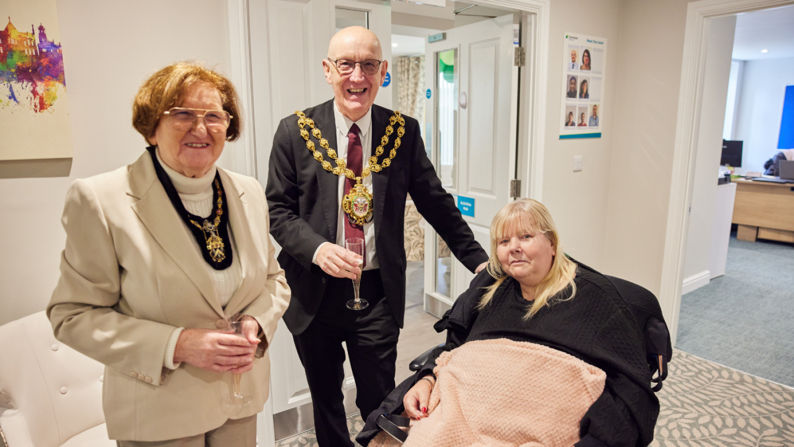 A man in a formal suit with a gold chain, and a woman in a beige suit, stand smiling beside a seated woman with a pink blanket, in an office setting.