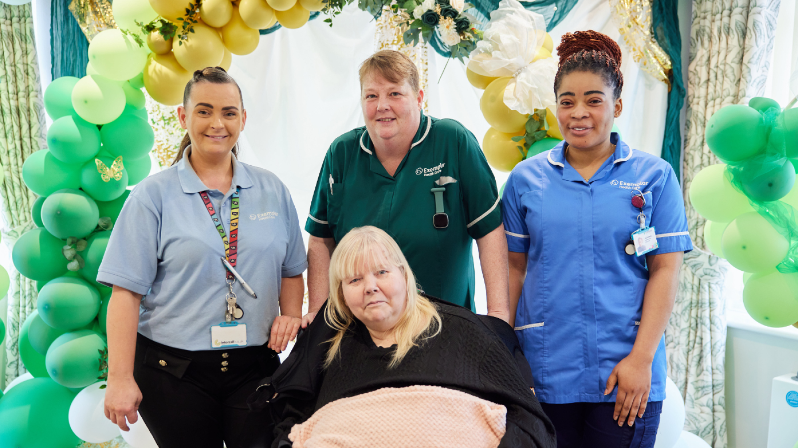 Four caregivers stand behind a seated woman covered with a blanket. They smile amid green and gold balloon decorations, conveying a warm, celebratory atmosphere.