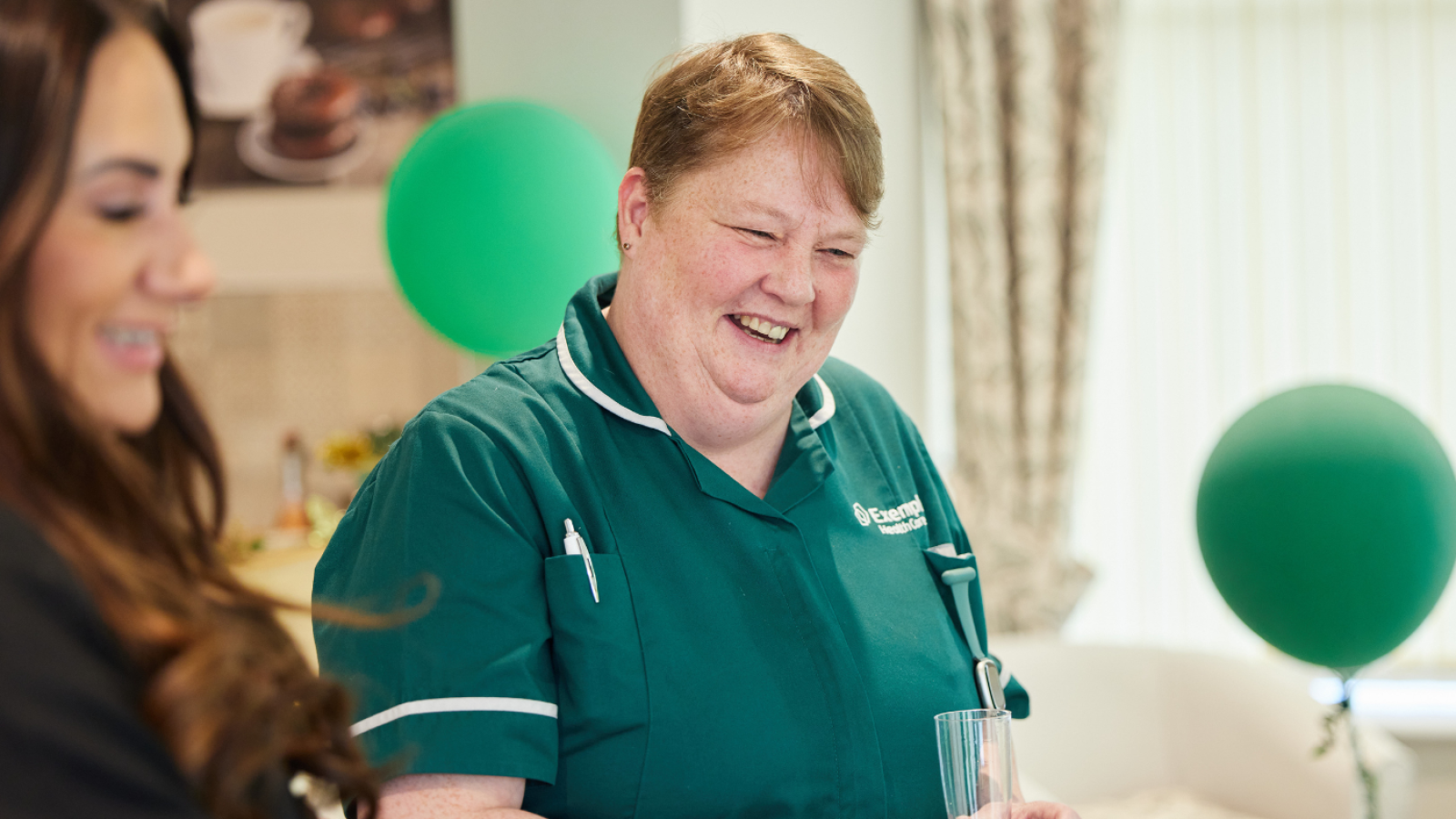 A smiling woman in a green uniform stands holding a glass in a cozy room with green balloons. Another person is beside her. The mood is cheerful.