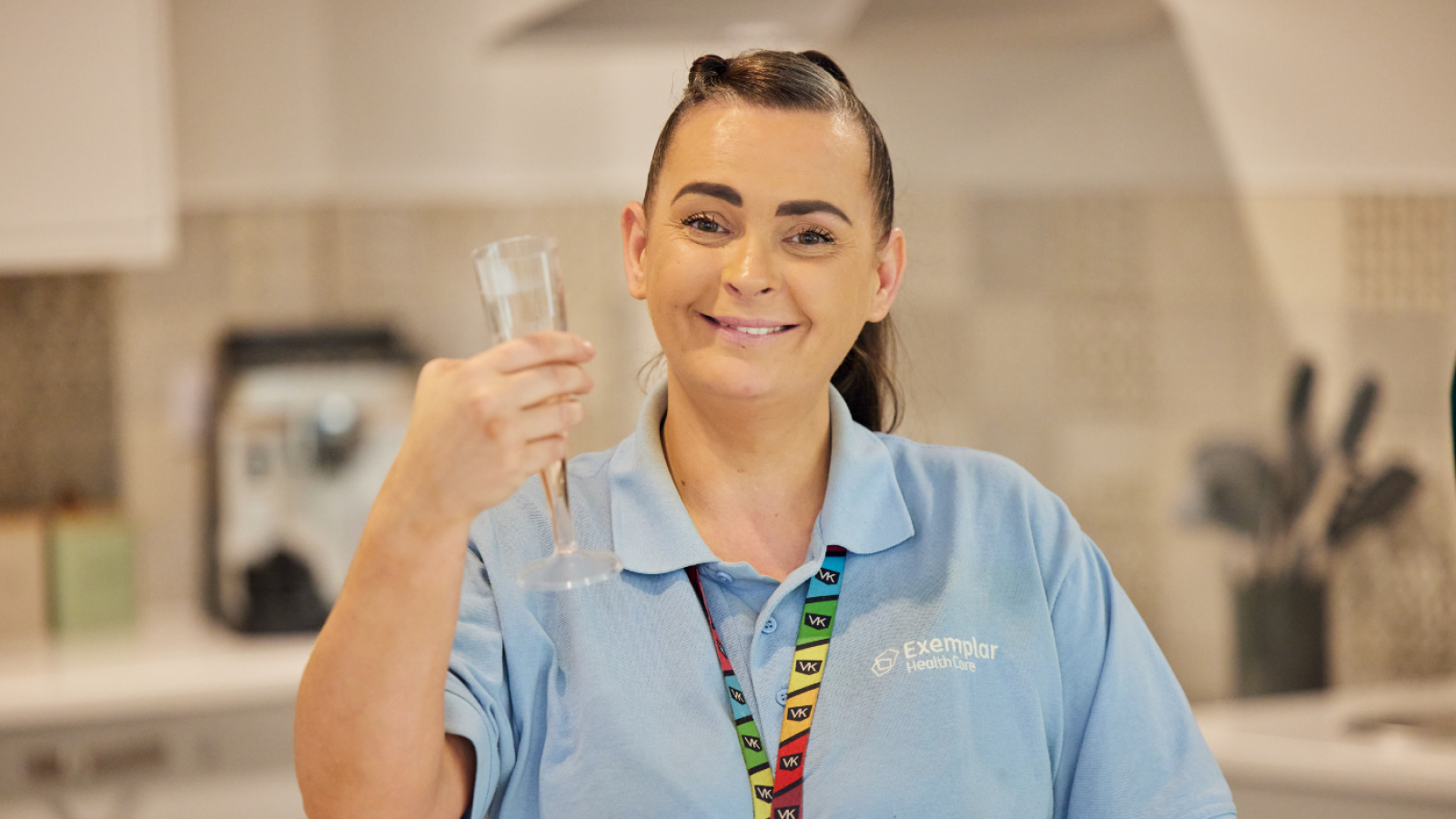 Smiling woman in a blue uniform holding an empty champagne flute in a kitchen. She's wearing a colorful lanyard and exudes a cheerful demeanor.