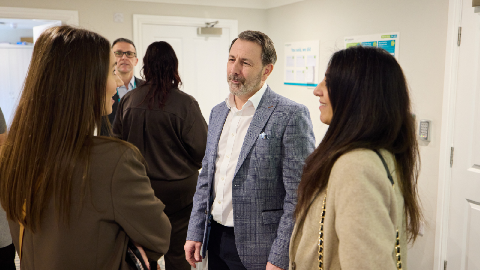 A group of professionally dressed people are engaged in conversation in a bright room. The atmosphere is friendly and attentive.
