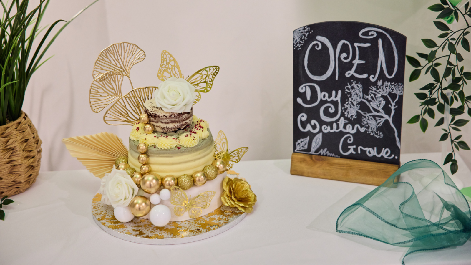 Innovative cake with gold accents, white roses, and leaf motifs next to a chalkboard reading "Open Day Walton Grove." Elegant and festive setting.