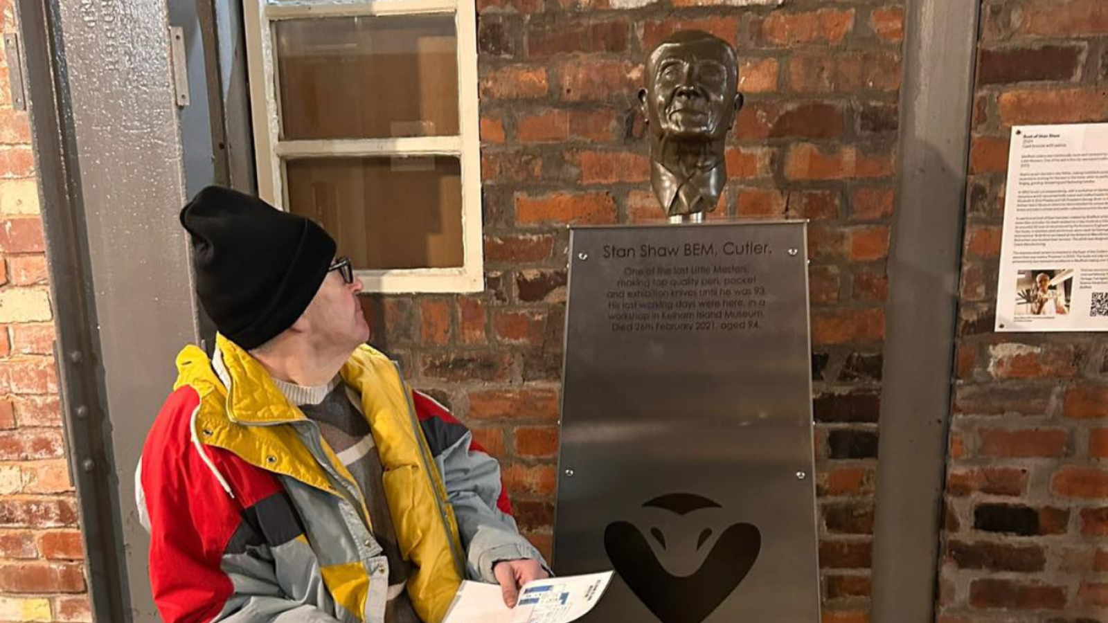A man wearing a jacket and hat is seated next to a statue, enjoying the peaceful atmosphere of the surroundings