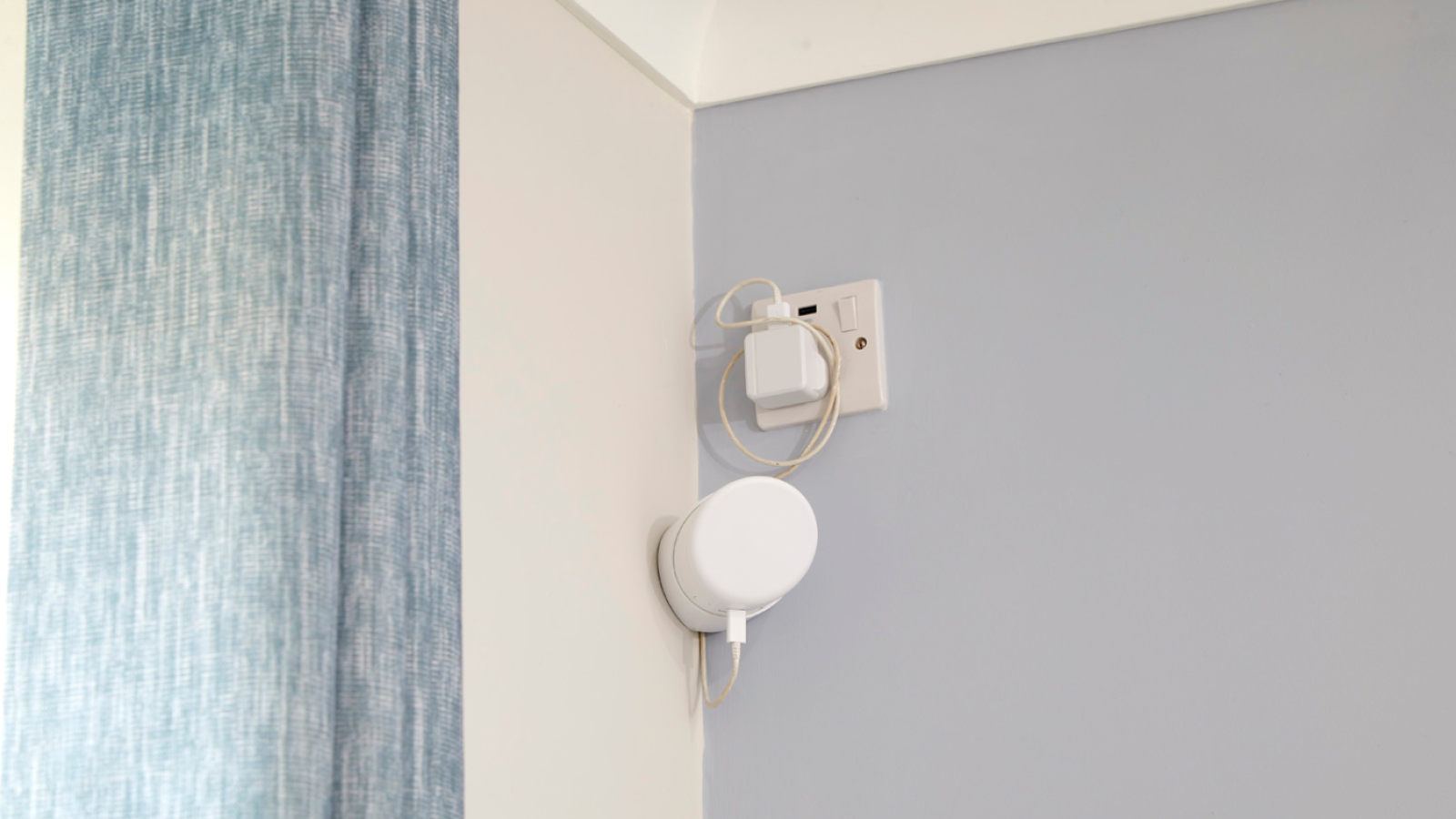 A white wireless device is mounted on a corner wall near a power outlet. Blue curtains hang on the left. The room has a calm, minimalistic feel.