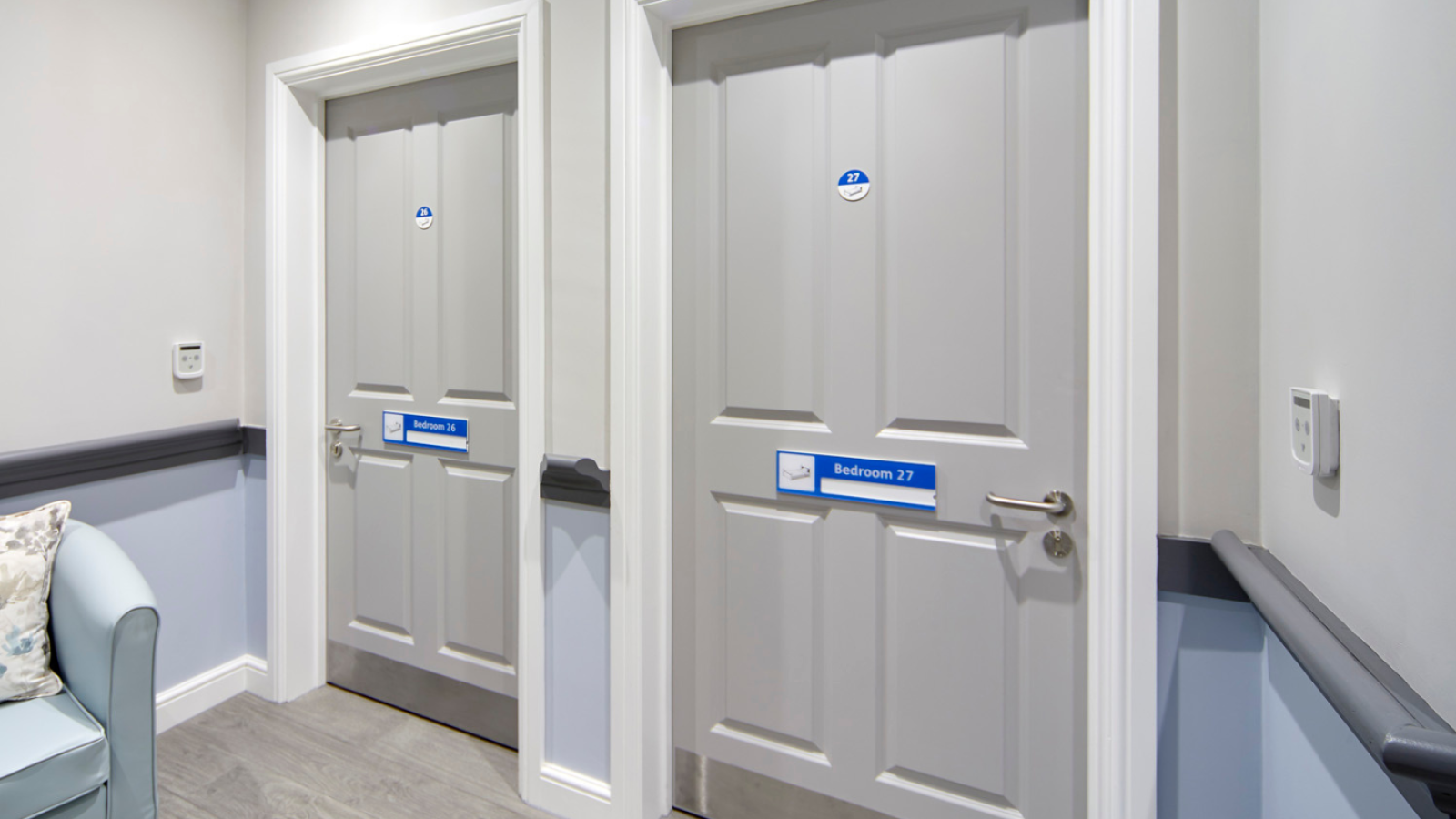 Gray corridor with two doors labeled "Bedroom 26" and "Bedroom 27" on blue plaques. A cushioned chair with pillows is partially visible on the left.