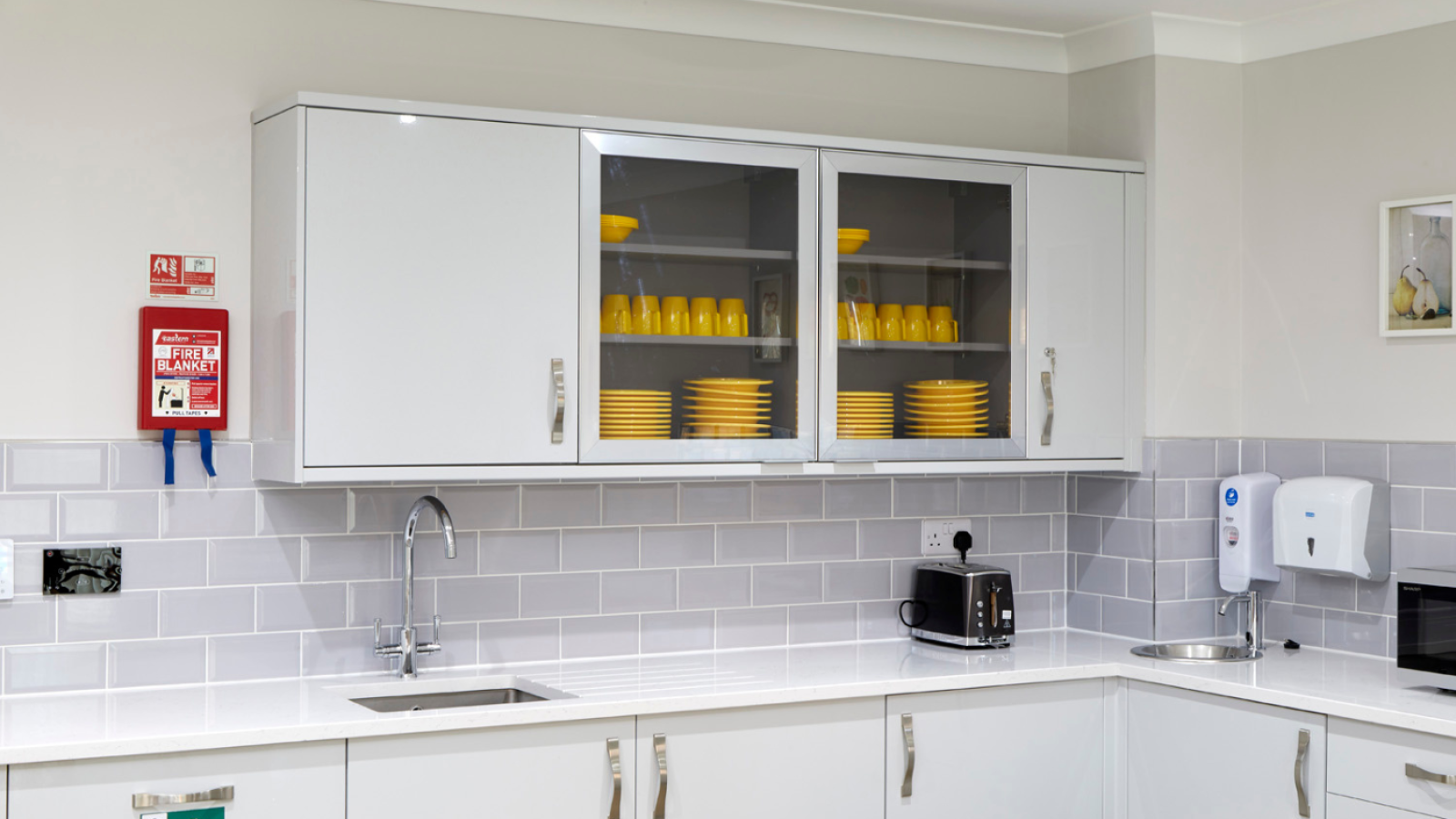 Modern kitchen with white cabinets and grey backsplash. Yellow dishes are displayed in glass cabinets. A toaster, fire blanket, and soap dispenser are on the counter.