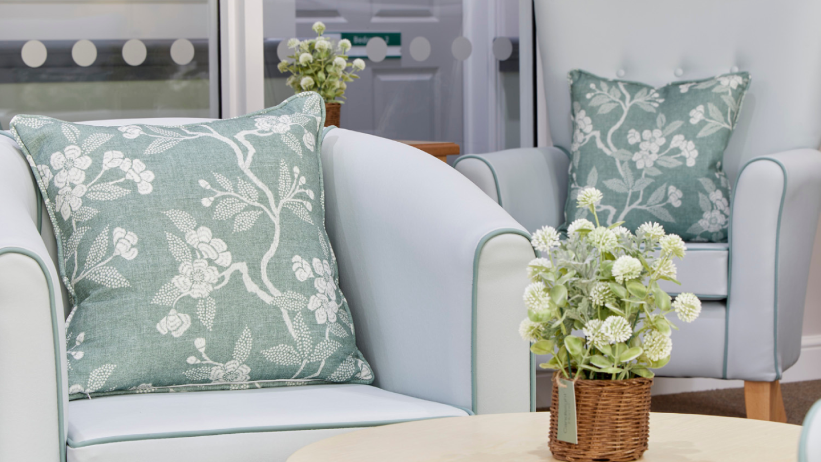Light blue armchairs with floral cushions are in a cozy room. A small round table with a basket of white flowers adds a welcoming touch.