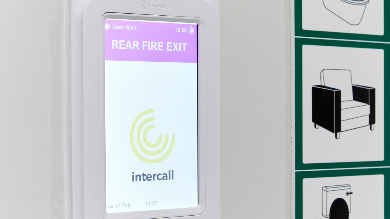 White wall-mounted digital display showing a "Rear Fire Exit" alert with a purple header and the Intercall logo. Icons of chair and toilet are nearby.