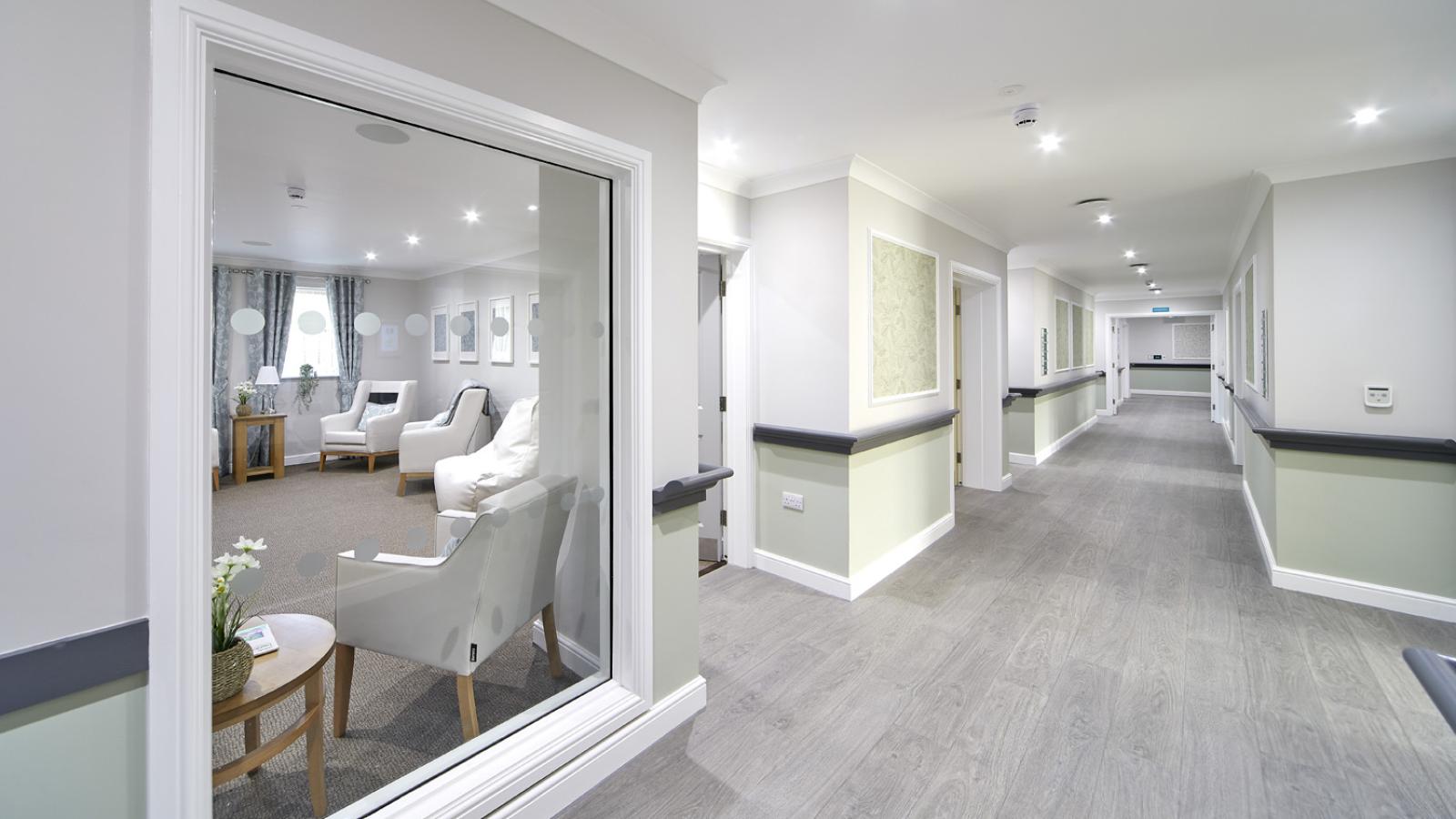 A bright, spacious corridor with light wood flooring and soft white walls. A window on the left shows a cozy lounge with armchairs and a small table.