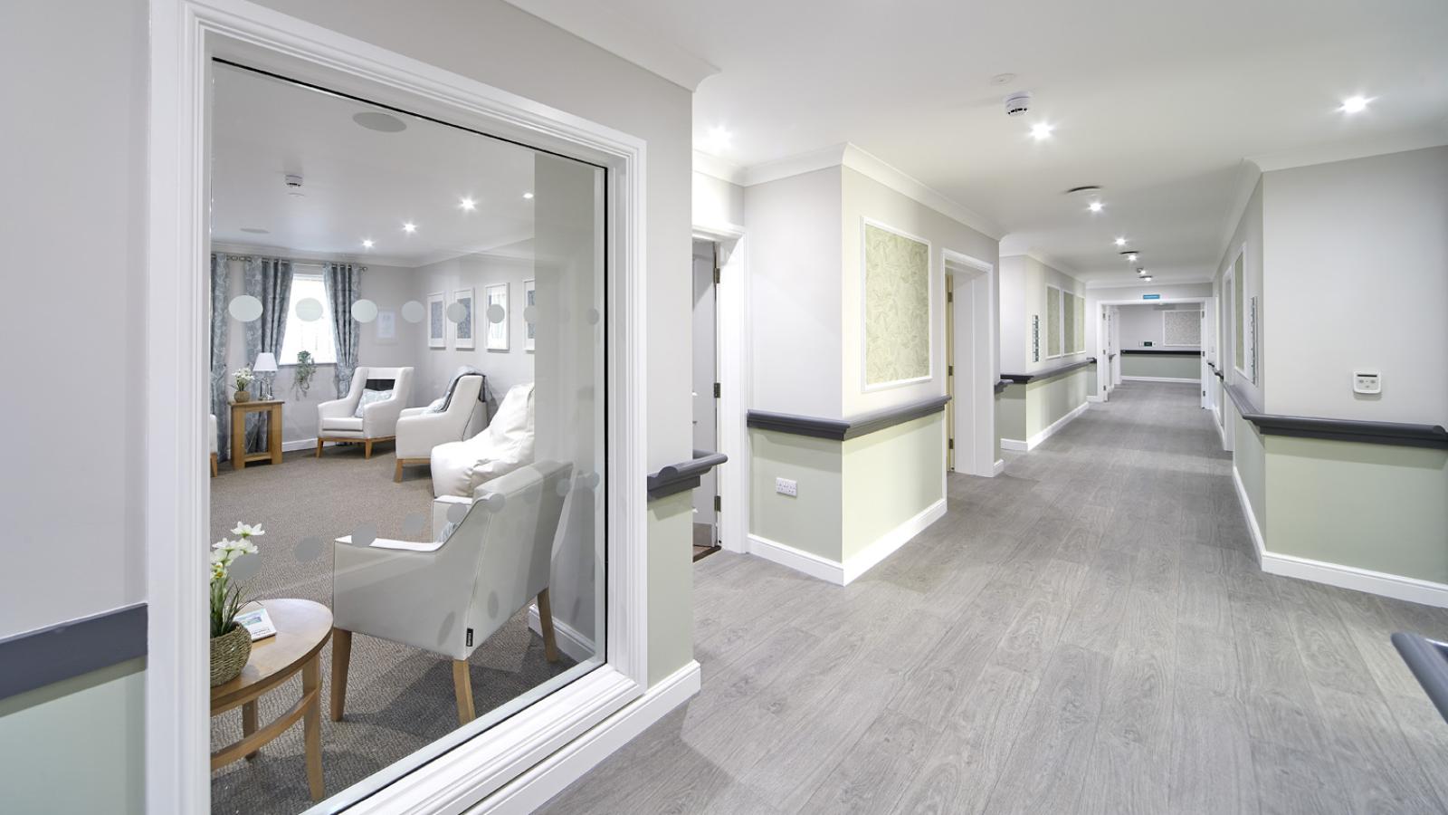 A bright, spacious corridor with light wood flooring and soft white walls. A window on the left shows a cozy lounge with armchairs and a small table.