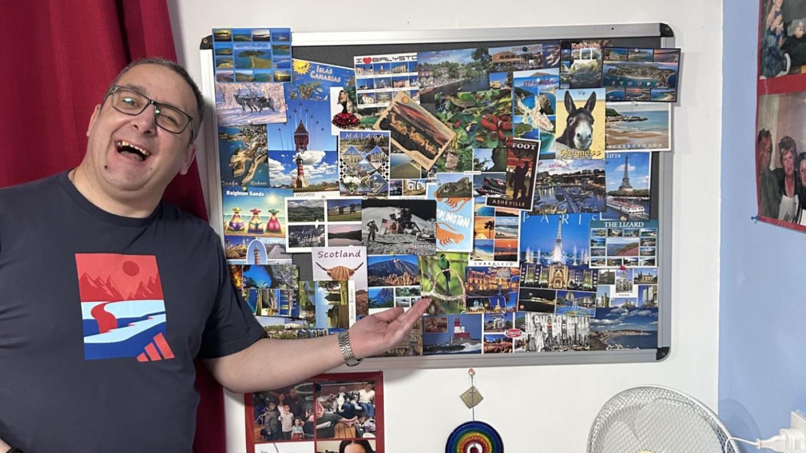 Man points to postcards displayed on the wall while smiling