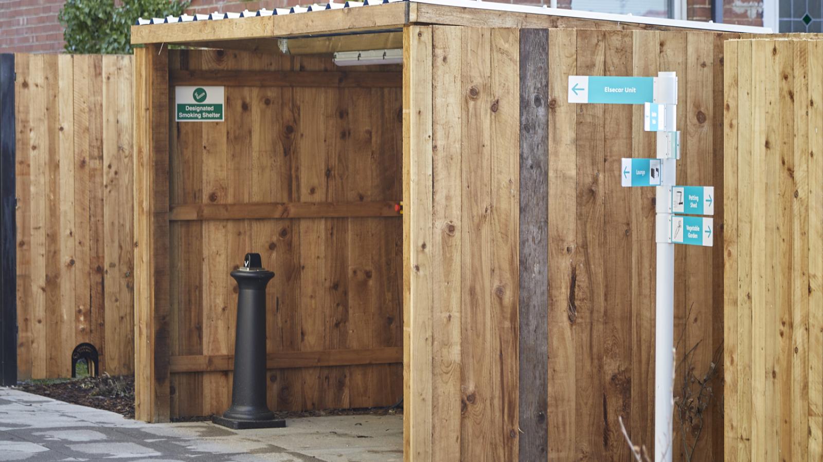 A wooden smoking shelter with a gray roof stands next to a tall signpost pointing to nearby units. A black ashtray sits inside on a paved area.