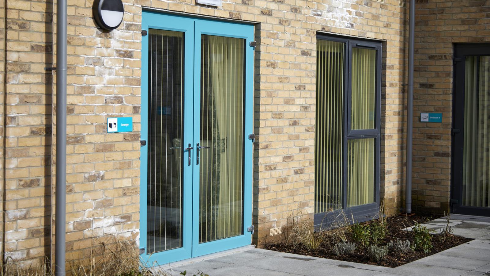 Brick building exterior with turquoise-framed double doors and large windows, both with vertical blinds. Small plants line the building’s base.