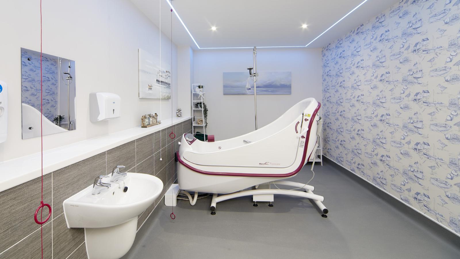 A clean, modern hospital bathroom featuring a sleek white bathtub, a sink, and patterned wallpaper. Red pull cords hang from the ceiling, ensuring safety.