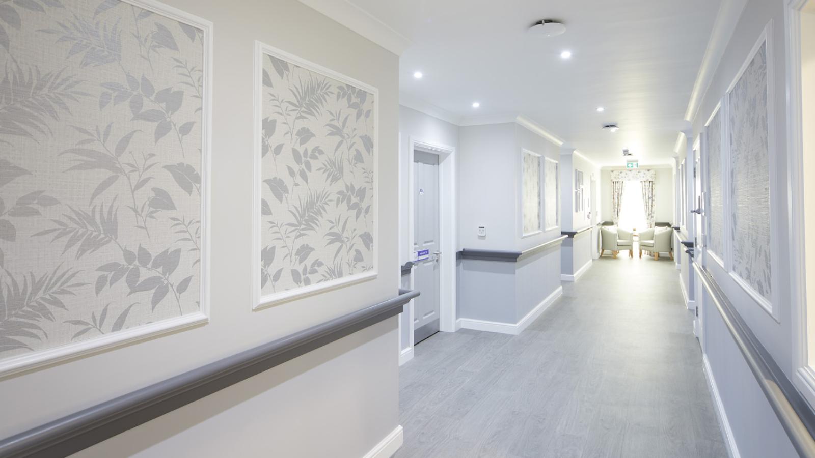 Bright, modern hallway with soft lighting, floral patterned wall panels, and light grey flooring. At the end are two chairs near a window, evoking a calm atmosphere.
