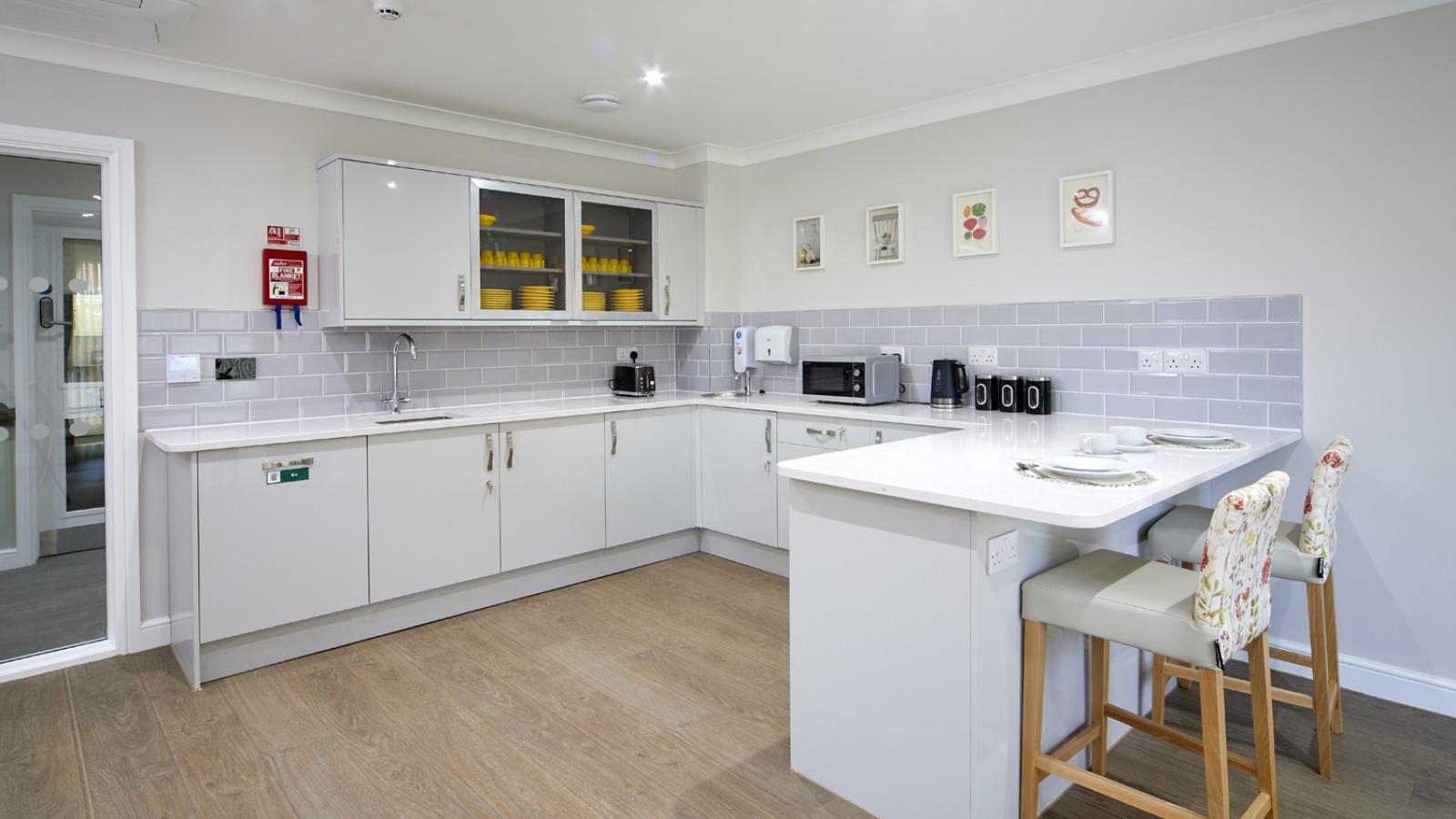 Modern kitchen with white cabinets, gray backsplash, and wood flooring. Yellow dishes in glass cabinets, a white counter with floral chairs, and small appliances create a cozy feel.