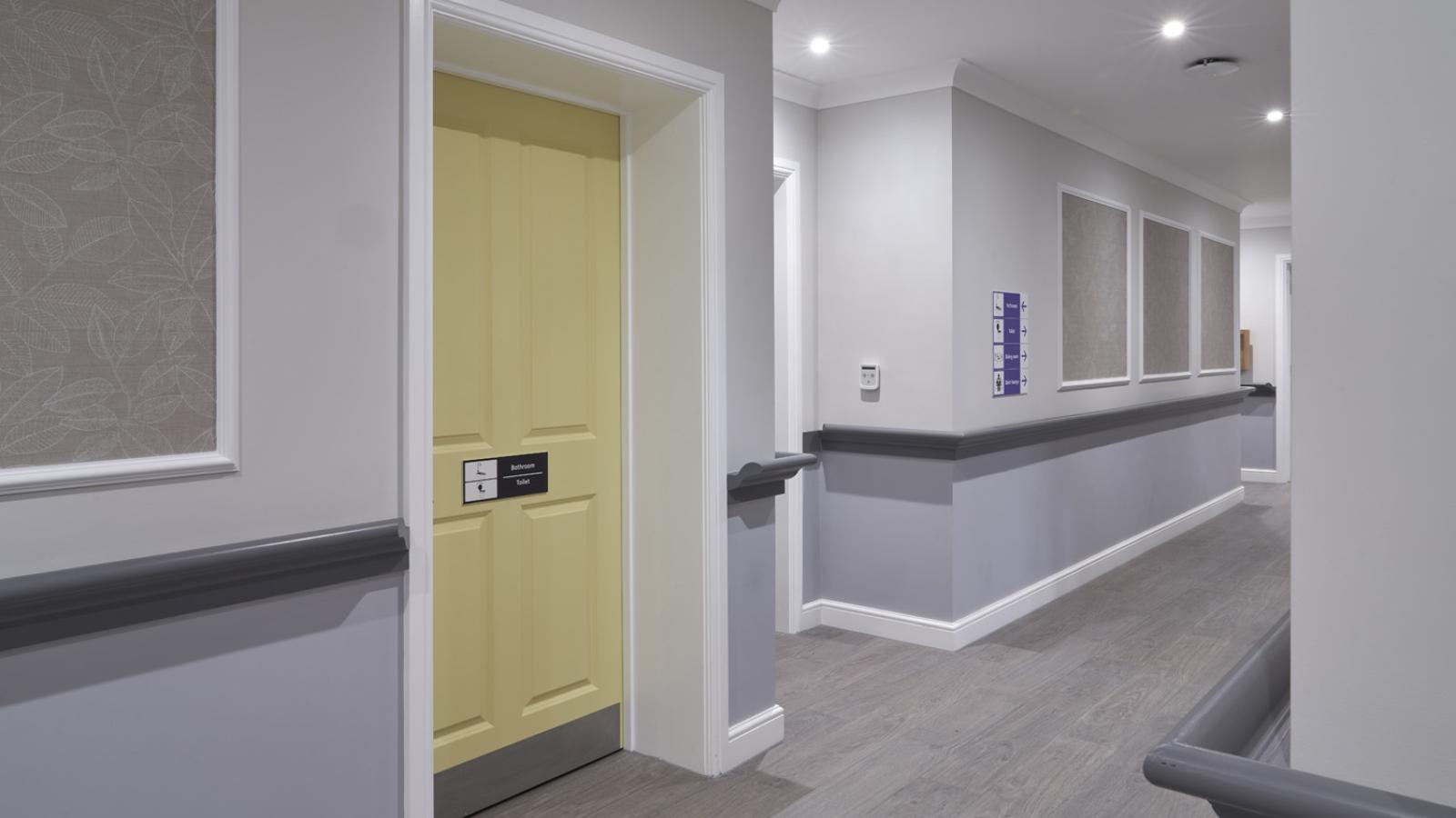 A modern hallway with light gray walls and a wooden floor. A yellow door with name plates is on the left. The hallway is well-lit and orderly.