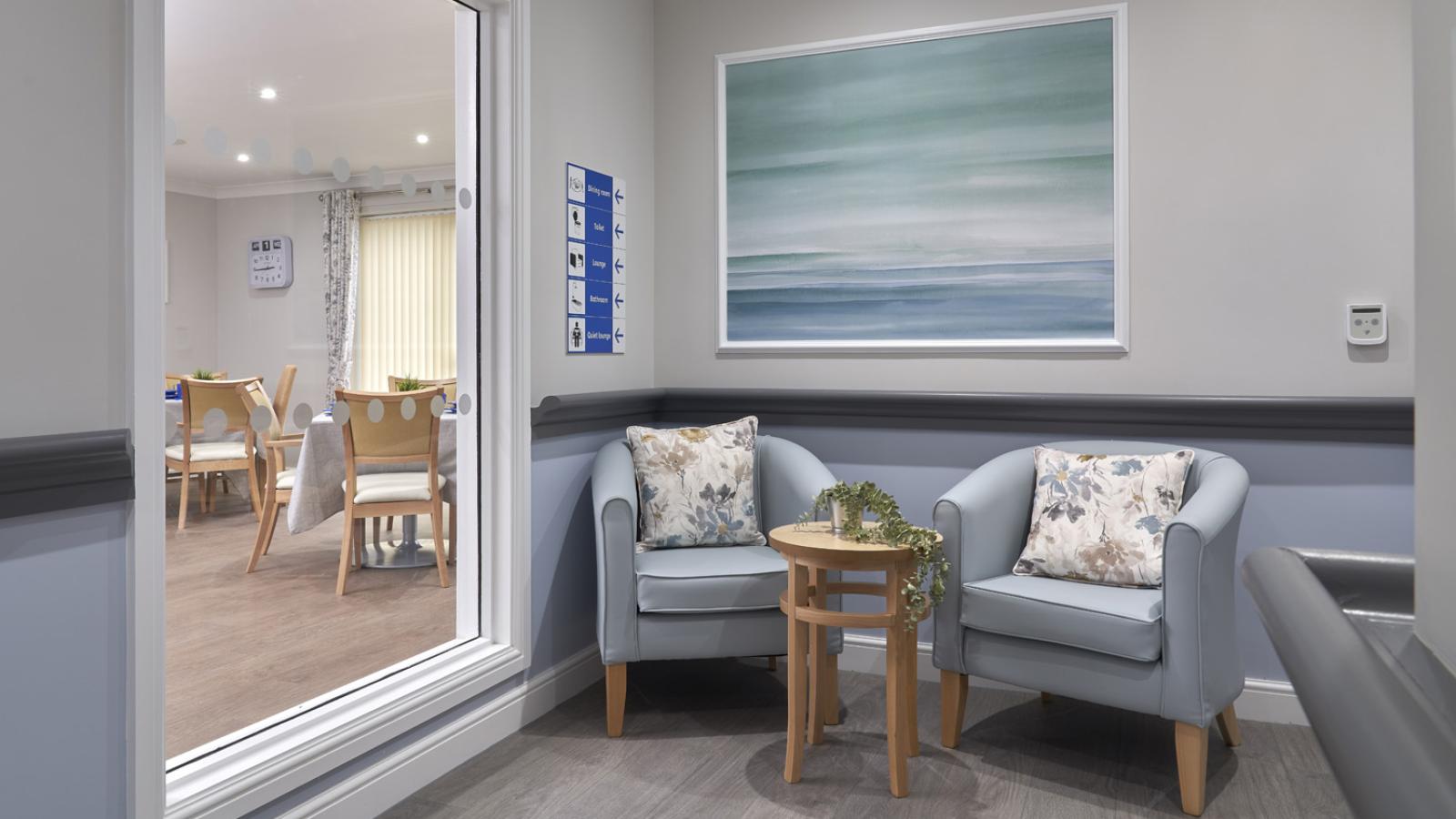Cozy seating area with two light blue armchairs and a small wooden table. A serene ocean painting hangs above. Adjacent room visible through window.