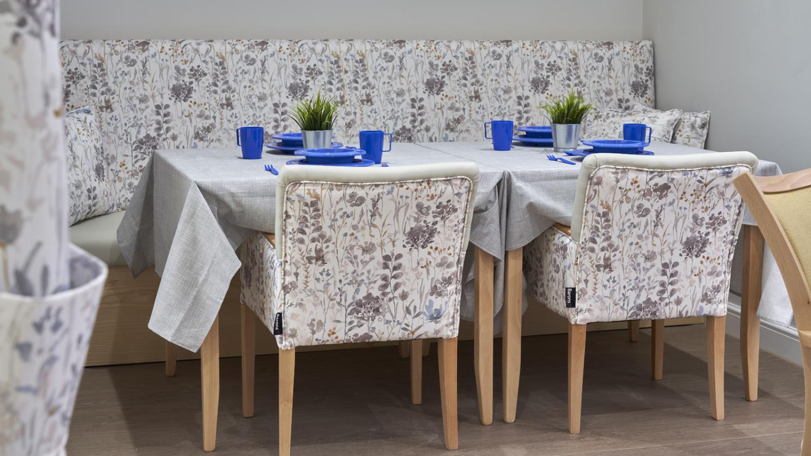 Cozy dining area with floral-patterned chairs and a matching bench, covered with light-gray tablecloths. Blue table settings and potted plants add contrast.
