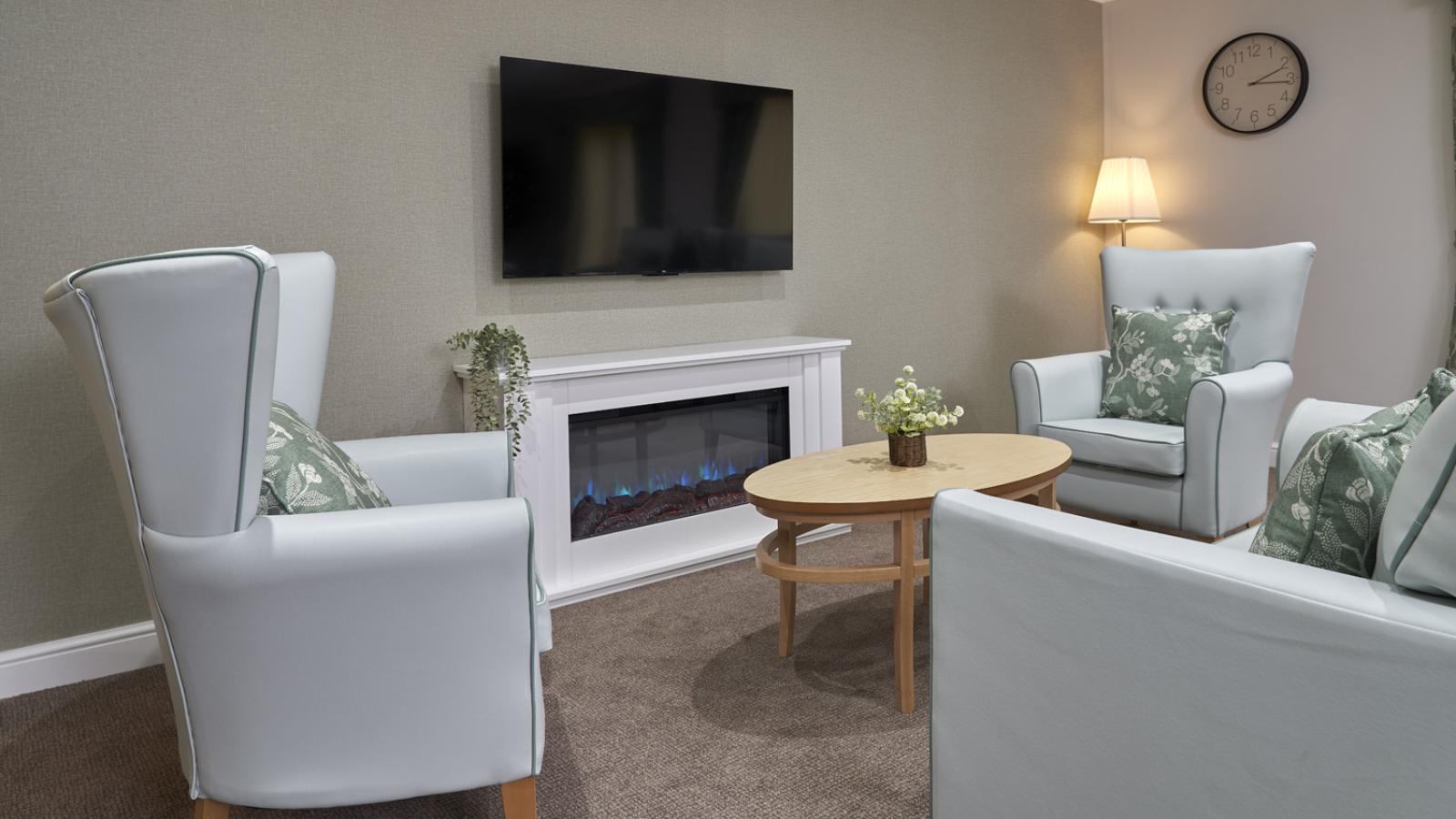A cozy living room with light blue armchairs, a small wooden table with flowers, a wall-mounted TV above an electric fireplace, and soft ambient lighting.