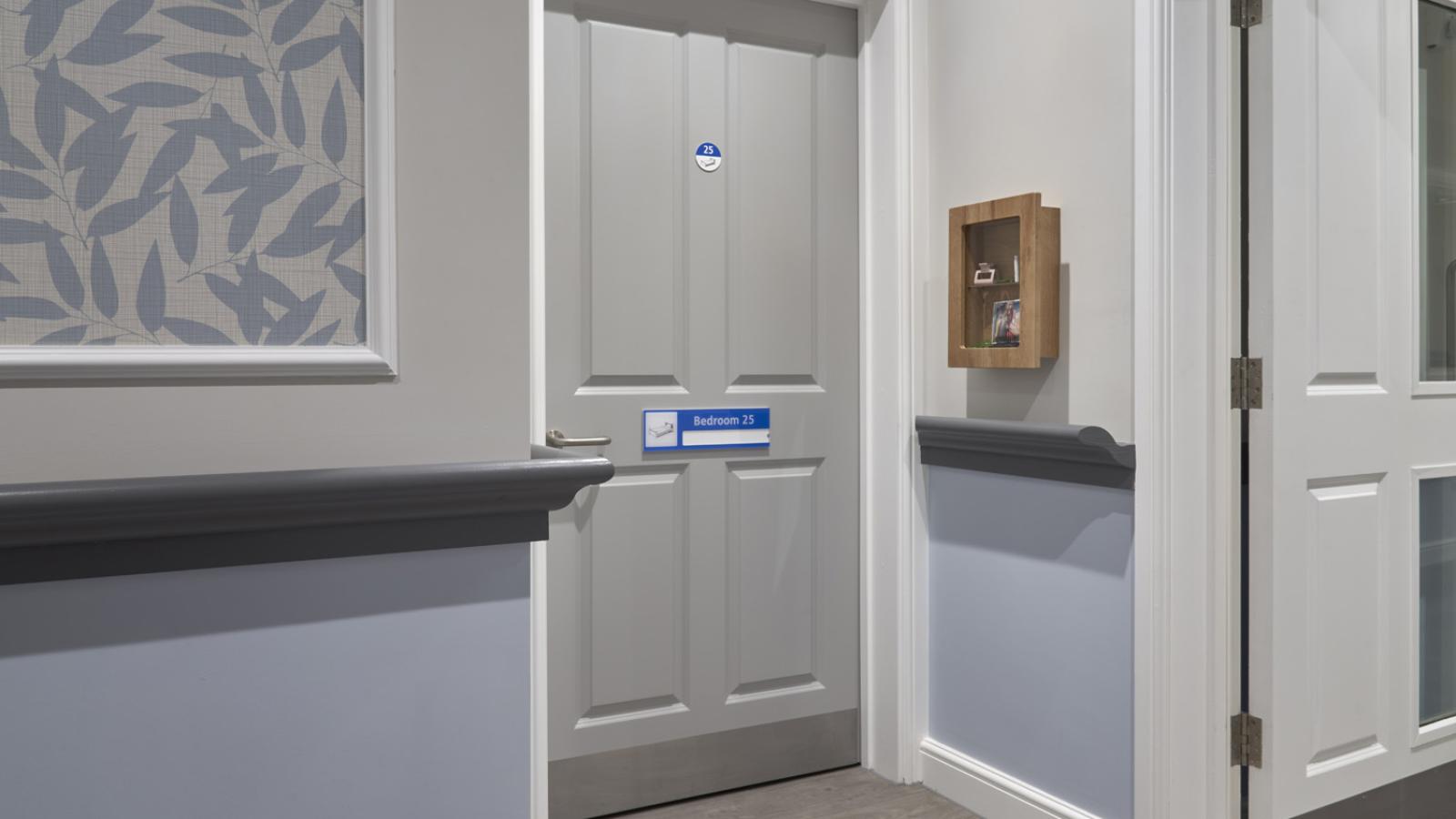 Gray door labeled "Bedroom 25" in a hallway with light blue walls and leaf-patterned wallpaper. A wooden box on the wall contains papers, creating a serene atmosphere.