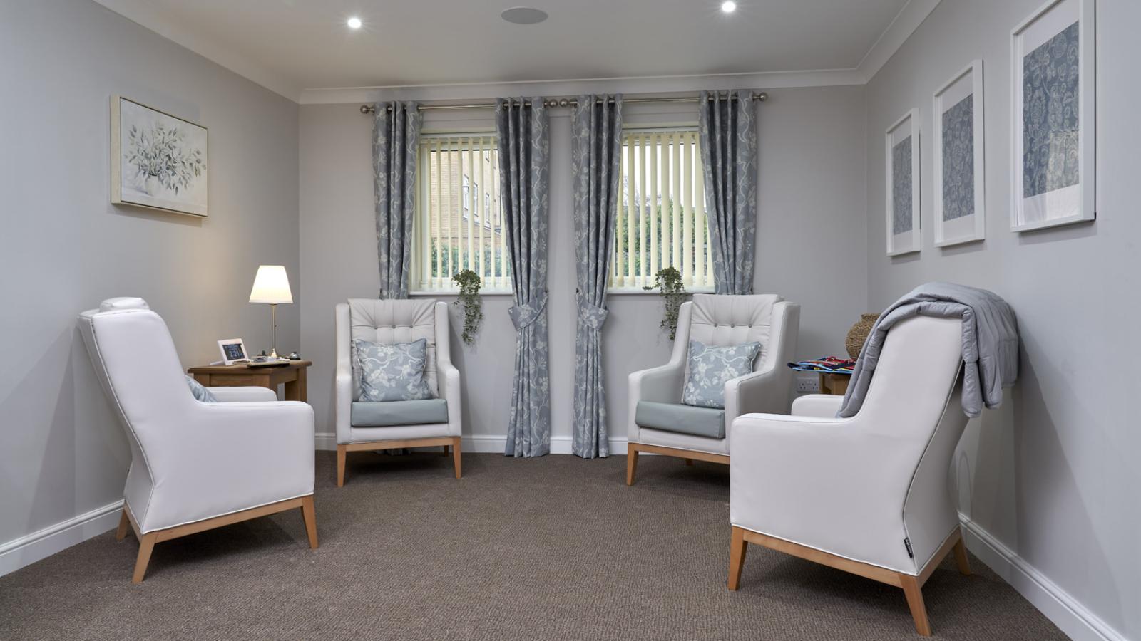 Cozy room with four white armchairs arranged in a circle on brown carpet. Two windows with blue curtains flank the space. Soft lighting and wall art create a calm ambiance.