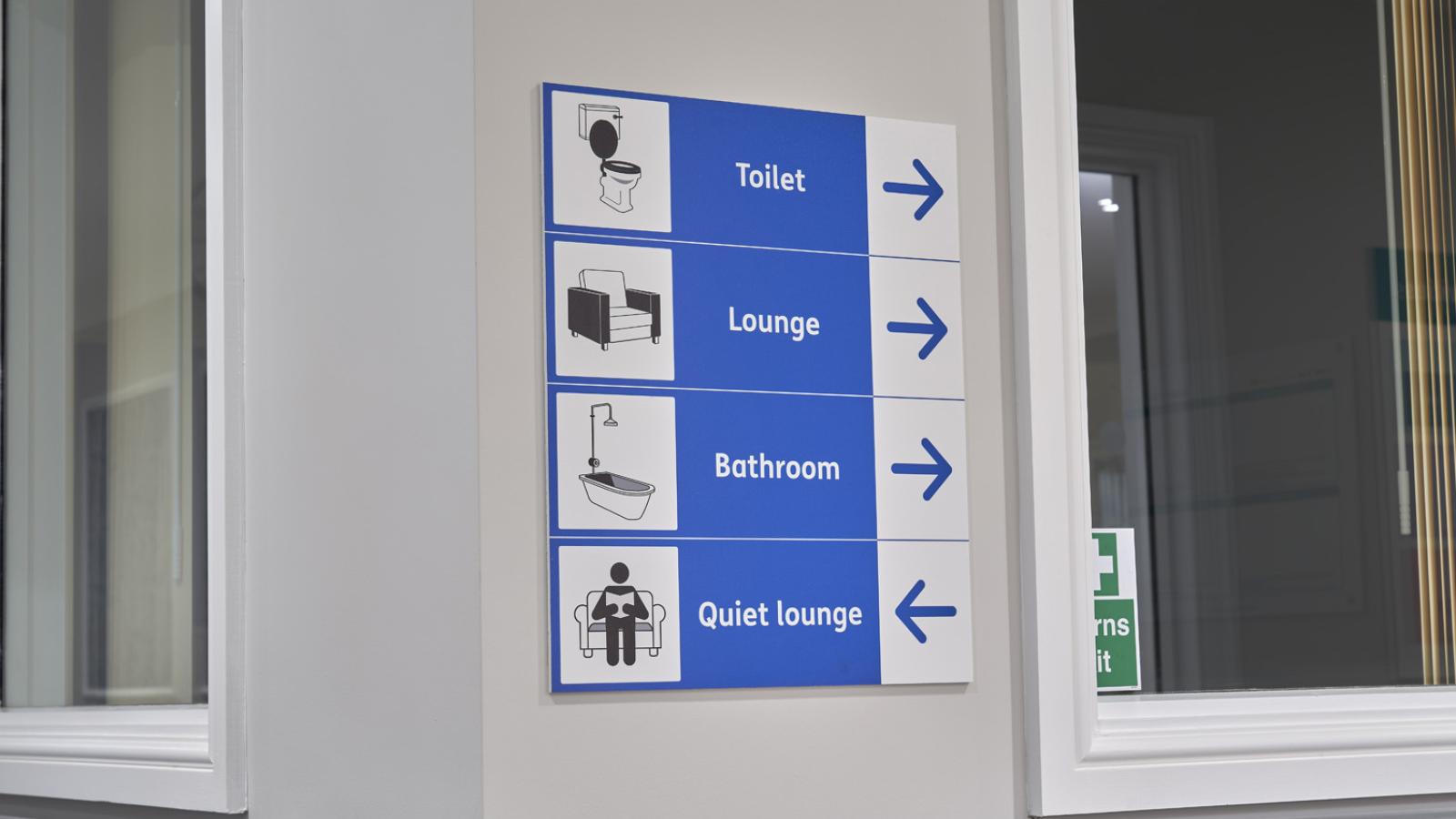 A wall sign with blue and white directional arrows and icons points to facilities: Toilet, Lounge, Bathroom to the right, Quiet Lounge to the left.