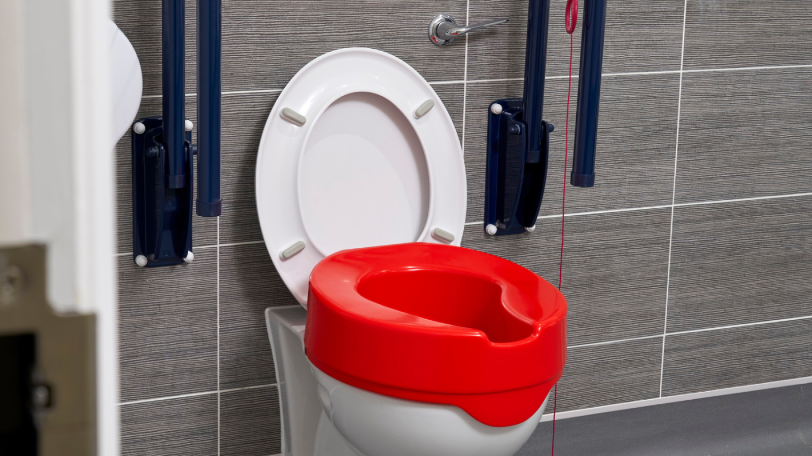 Accessible bathroom with a white toilet featuring a vibrant red raised seat, safety rails, and a pull cord against gray tiled walls. Functional and inclusive design.