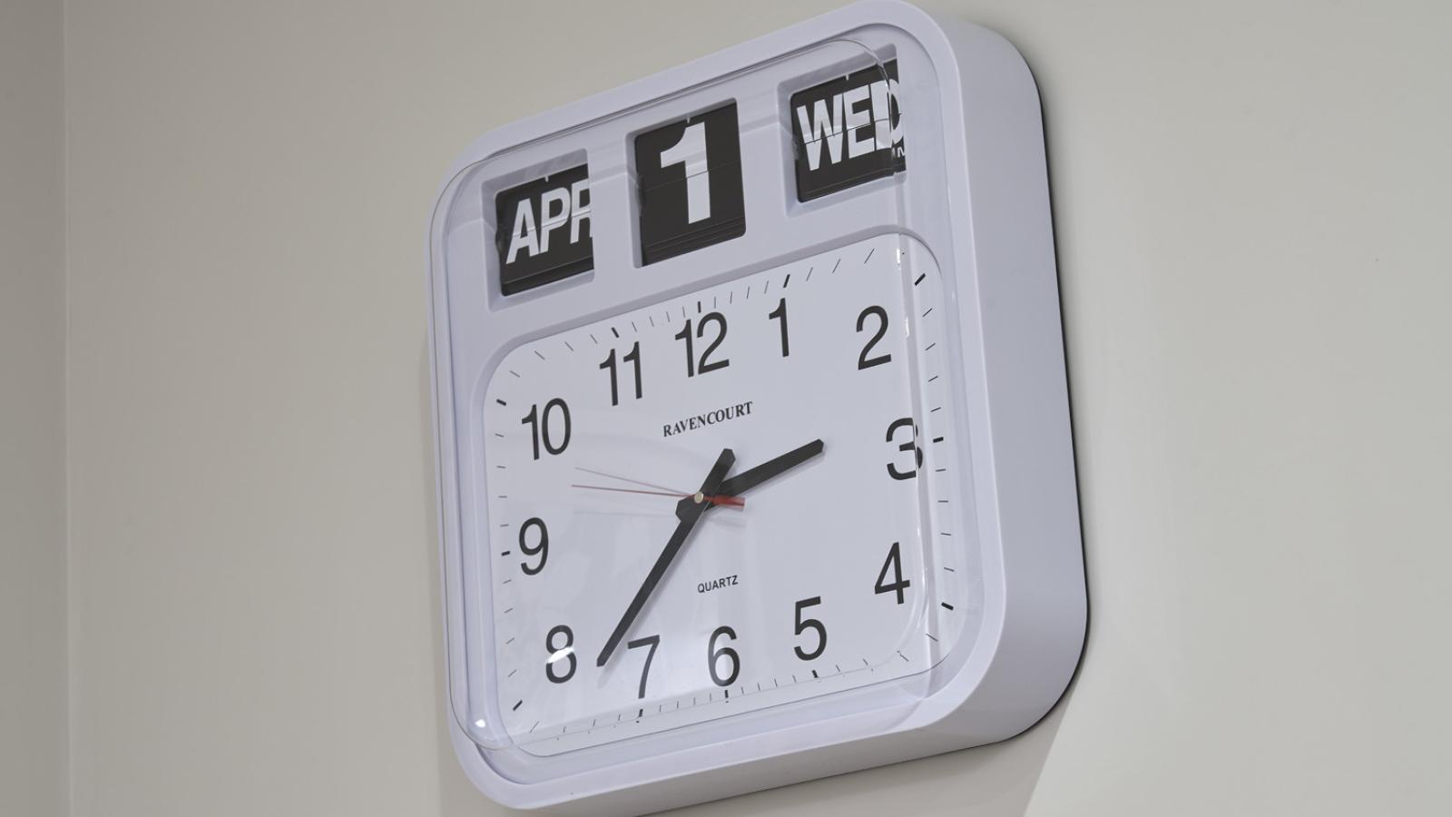 Square wall clock with a white face and black numbers displays 10:09. Above, flip-style panels show "APR 1 WED." The tone is modern and precise.