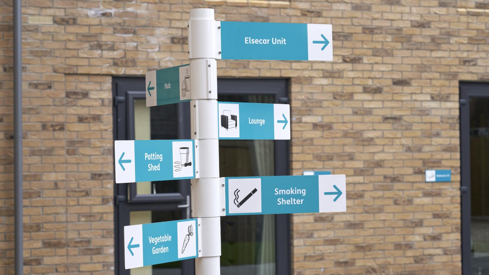 A signpost with blue and white directional arrows points to areas like Elsecar Unit, Lounge, Vegetable Garden, and Smoking Shelter against a brick building.
