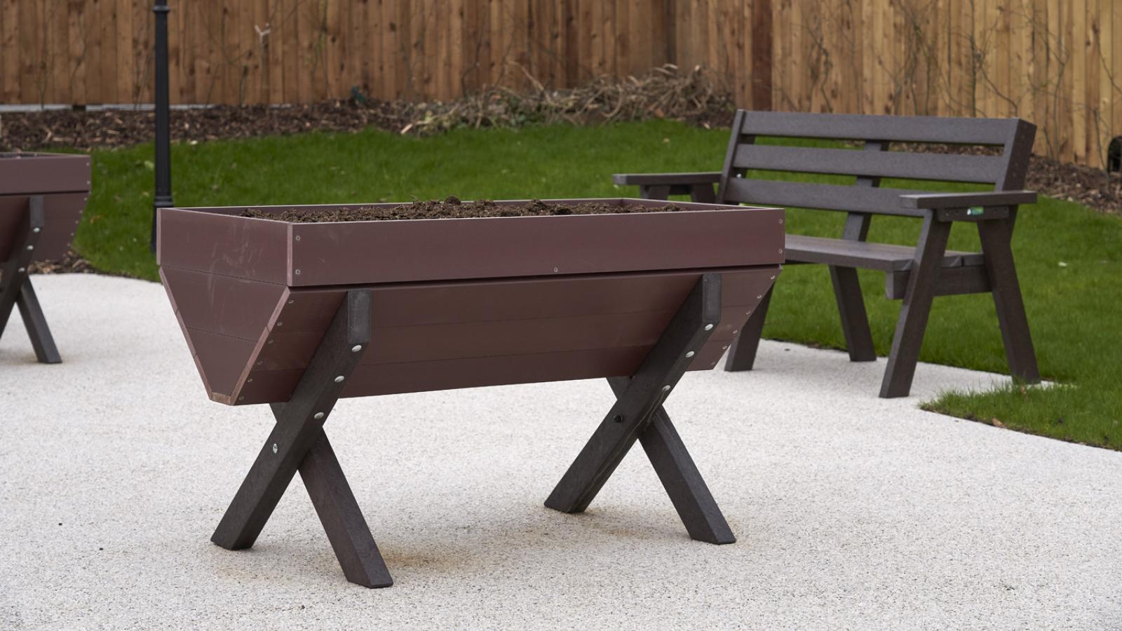 A brown, empty wooden planter on a patio with a wooden bench, green grass, and a wooden fence in the background. A vintage lamppost adds a peaceful ambiance.