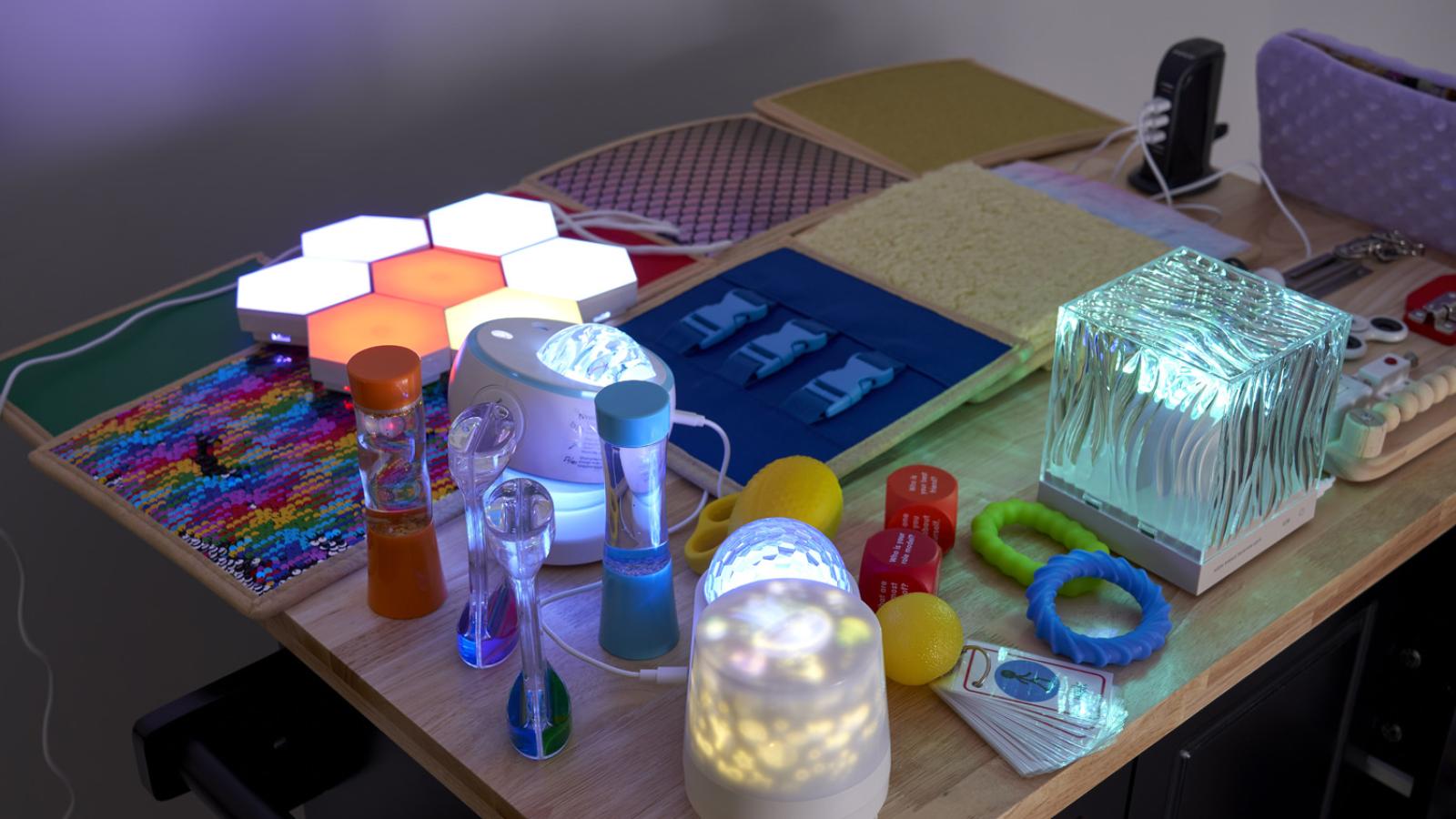 A wooden table filled with sensory toys: glowing hexagonal lights, liquid timers, textured pads, fidget items, and illuminated objects, creating a playful and calming atmosphere.